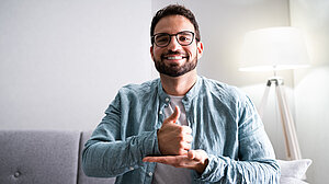 Mann mit Brille sitzt lächelnd auf Sofa und zeigt einen Daumen hoch